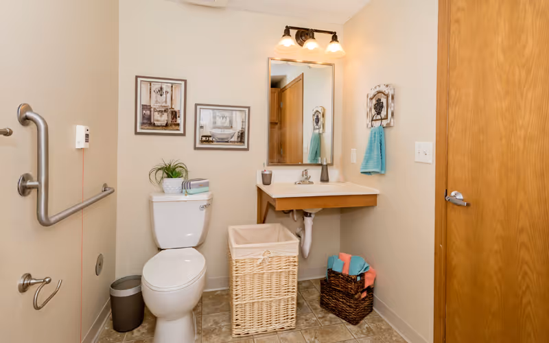 Accessible bathroom with a toilet, grab bars, wall-mounted sink and mirror, and storage baskets with towels.