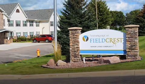 Entrance sign for Vista Prairie at Fieldcrest Senior Living Community with a large white building and parked red car in the background, surrounded by green grass and trees under a partly cloudy sky.