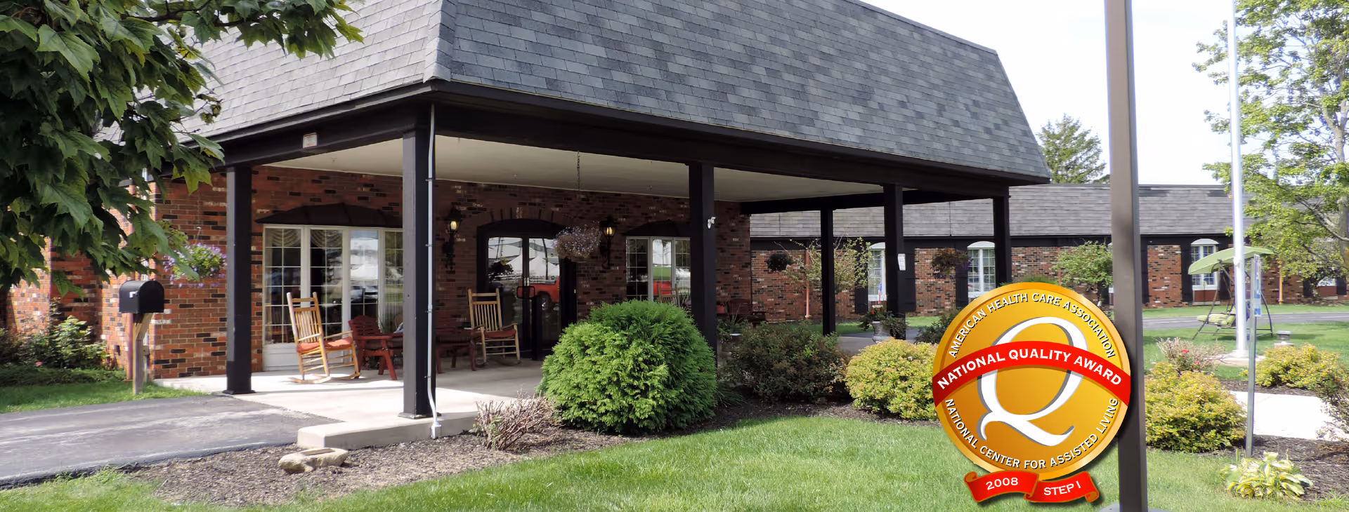 Exterior view of Van Wert Manor, a brick building with a covered entrance supported by black pillars. There are rocking chairs on the porch and well-maintained landscaping with green grass, bushes, and trees. A National Quality Award emblem is displayed in the lower right corner.