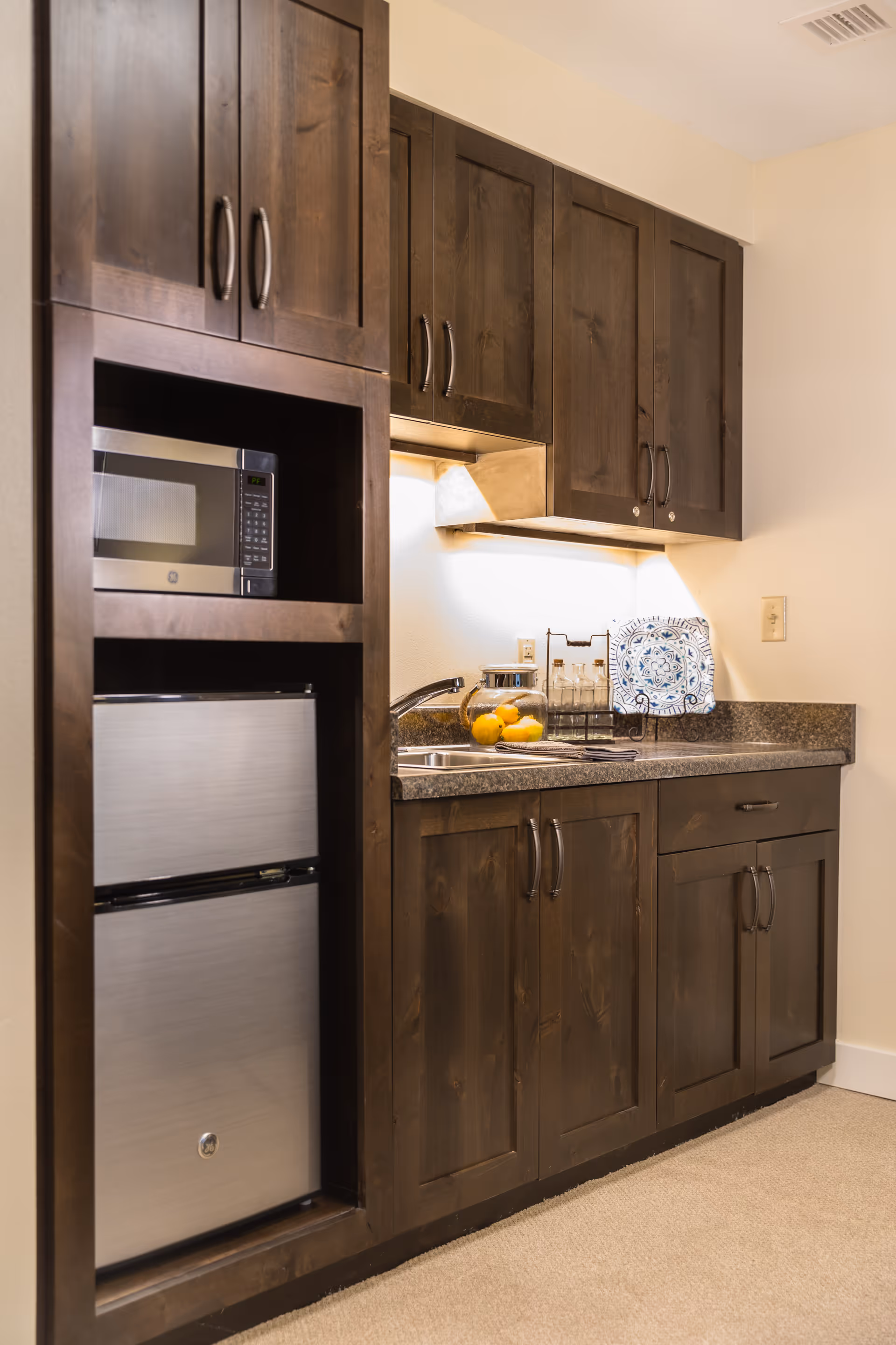 A kitchenette area with dark wooden cabinets, a small stainless steel refrigerator, a microwave, a sink, and a countertop with decorative items including a glass jar with lemons, bottles, and a decorative plate.
