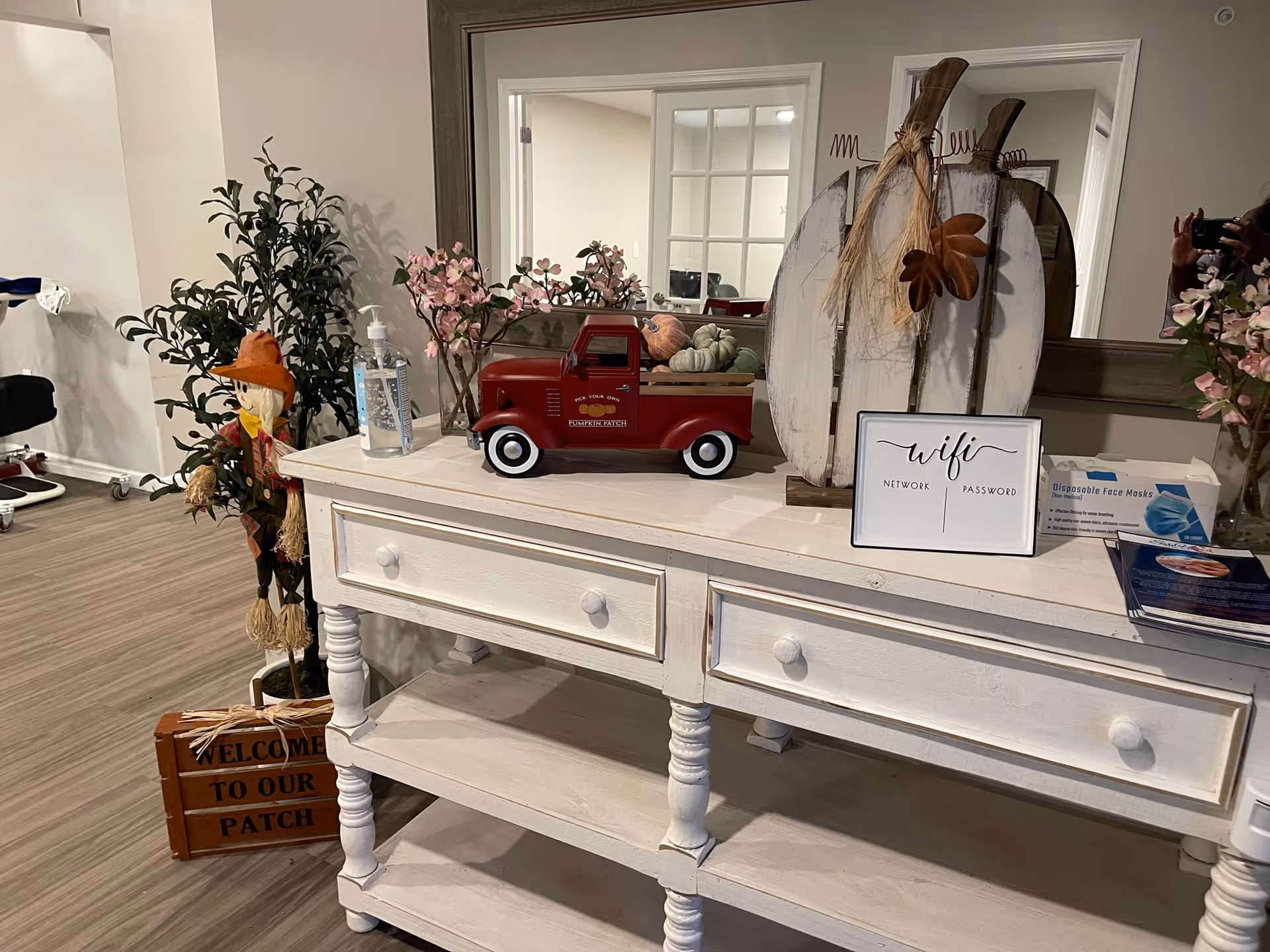 A white wooden console table with two drawers and a lower shelf, decorated with a small red toy truck carrying miniature pumpkins, a hand sanitizer bottle, a decorative sign for wifi network and password, and a rustic wooden pumpkin decoration. To the left of the table is a small scarecrow figure and a wooden crate sign that says 'Welcome to our patch'. The background shows a large mirror and part of a room with light wood flooring.