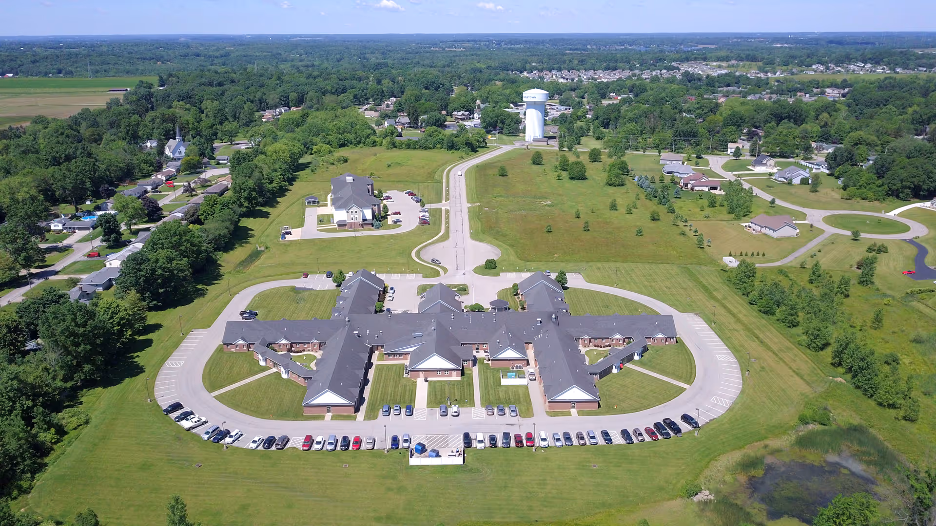 Aerial view of Masternick Memorial facility surrounded by green lawns and trees, with a large parking lot filled with cars in front. The building has a cross-shaped layout with multiple wings and a main entrance. Residential houses and a water tower are visible in the background under a clear blue sky.