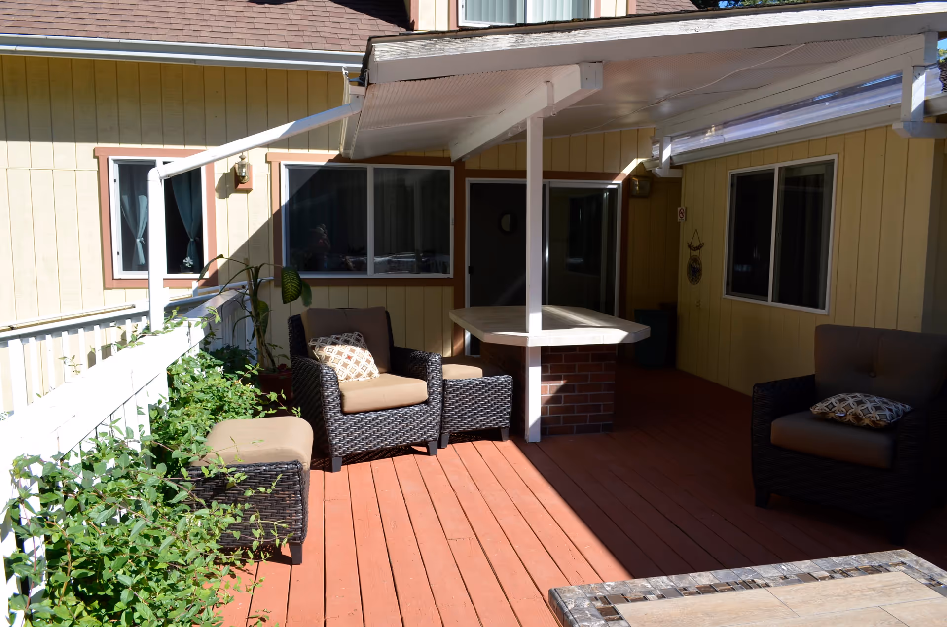 Outdoor patio area with wooden deck flooring, two cushioned wicker chairs with patterned pillows, a cushioned wicker ottoman, a brick and wood bar counter under a white awning, and plants along a white railing. The patio is adjacent to a yellow building with multiple windows.