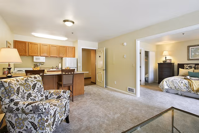 Interior view of a senior living apartment at Brookdale North Boulder featuring a living area with a patterned armchair and a glass coffee table, an open kitchen with wooden cabinets, a countertop with two chairs, and a bedroom visible through an open doorway with a bed, dresser, and lamp.