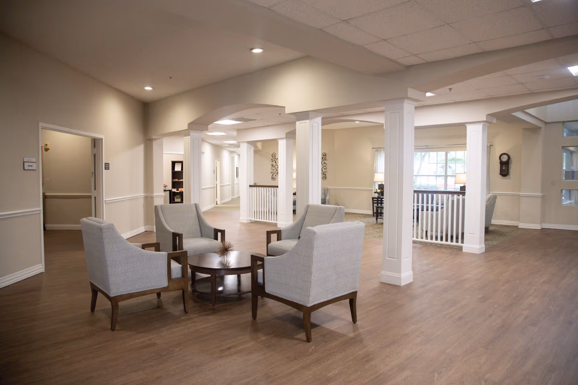 A spacious and well-lit common area in a senior living facility with four gray upholstered armchairs arranged around a round wooden coffee table. The room features light-colored walls, wooden flooring, white columns, and a large window letting in natural light. There is a hallway and additional seating areas visible in the background.