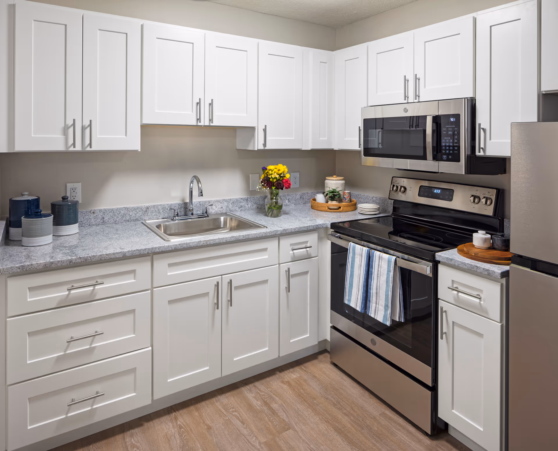 Modern kitchen with white cabinets, a stainless steel sink, stove, microwave, and refrigerator. The countertop is gray with a vase of colorful flowers and a tray with kitchen items. The floor is wood-style.