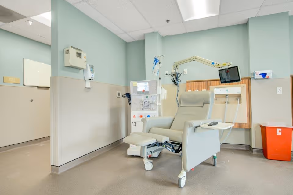 A medical treatment room with a reclining chair on wheels, a dialysis machine, a wall-mounted monitor, and medical equipment attached to the wall. The walls are painted light green and beige, and there is a red biohazard waste bin in the corner.