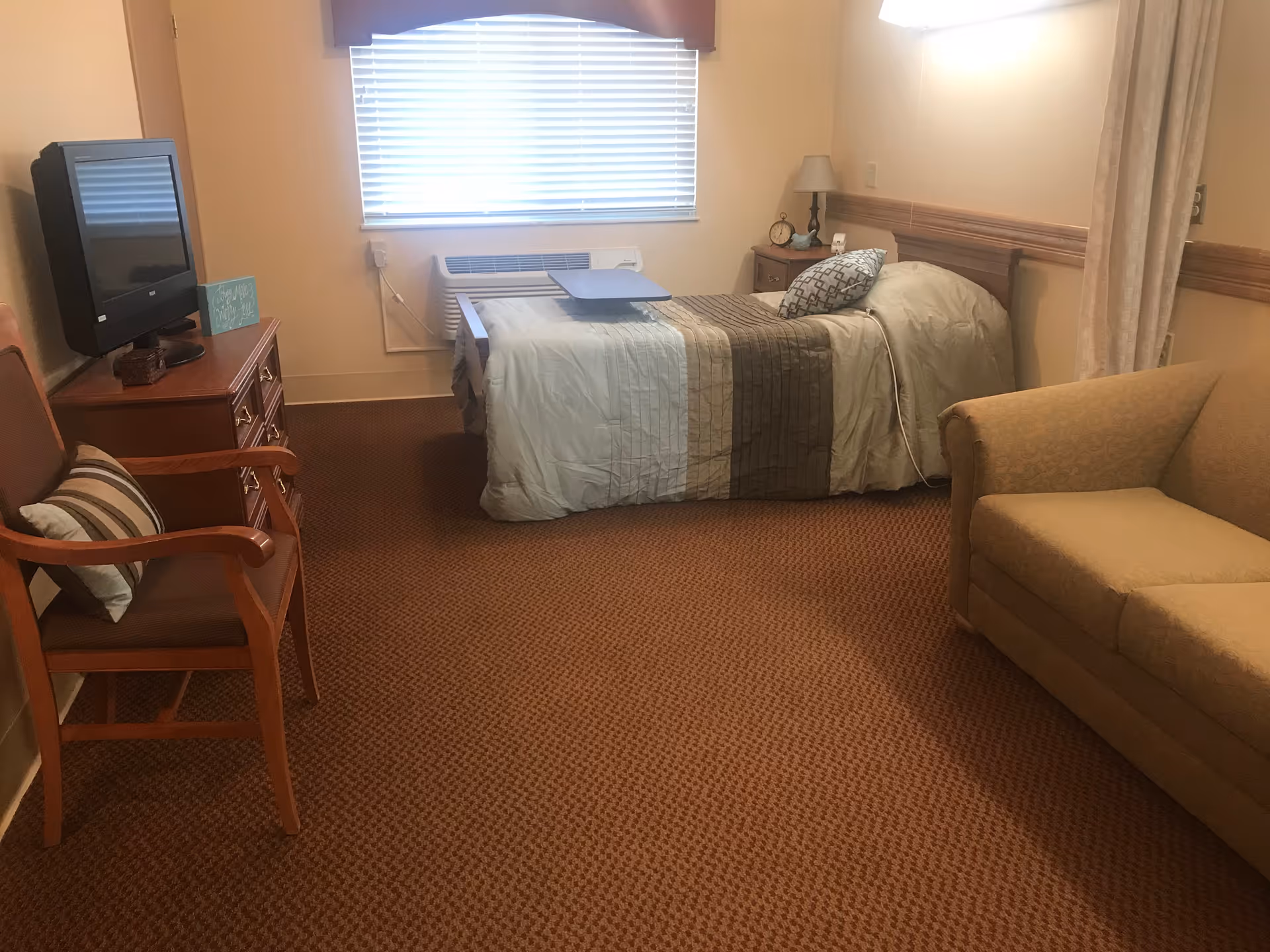 A senior living apartment room with a single bed covered in a striped comforter, a small bedside table with a lamp and clock, a window with blinds, a TV on a wooden dresser, a wooden chair with a cushion, and a beige couch.