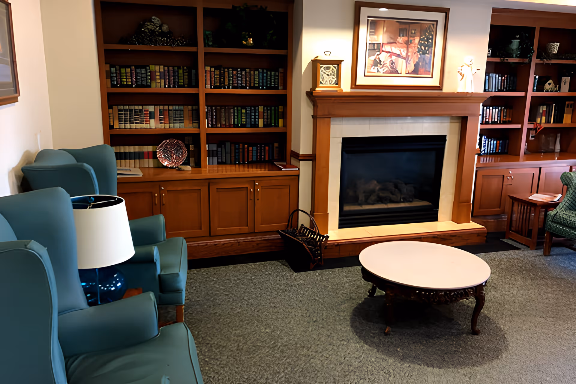 A cozy living room area with blue upholstered armchairs, a round wooden coffee table, a fireplace with a wooden mantel, and built-in wooden bookshelves filled with books and decorative items.