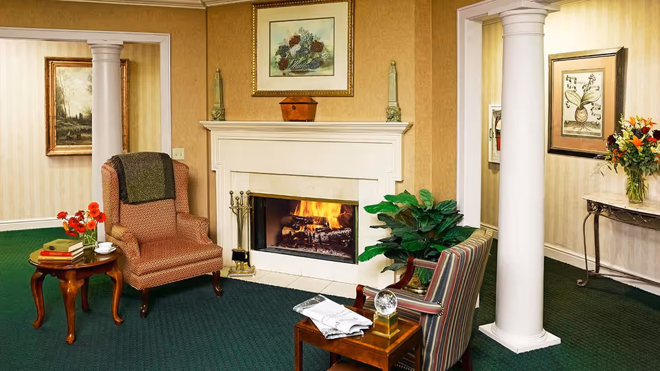 A cozy living room area with a lit fireplace in the center. There is a patterned armchair with a dark throw draped over it next to a small wooden side table holding a vase with red flowers, a cup, and some books. Across from the fireplace is a striped armchair with a small wooden table in front of it, holding a folded newspaper and a decorative glass orb. The room features green carpet, beige wallpaper, two white columns, framed artwork on the walls, and a potted plant near the fireplace.