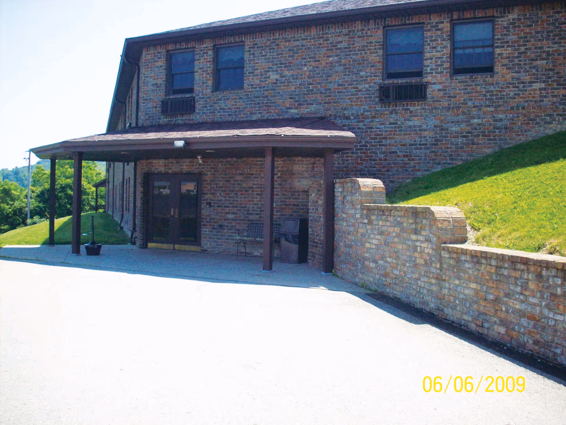 Exterior view of a brick building with a covered entrance supported by wooden posts. There is a bench and a trash can near the entrance. The building is situated on a slight hill with green grass surrounding it. The date 06/06/2009 is visible in the bottom right corner.