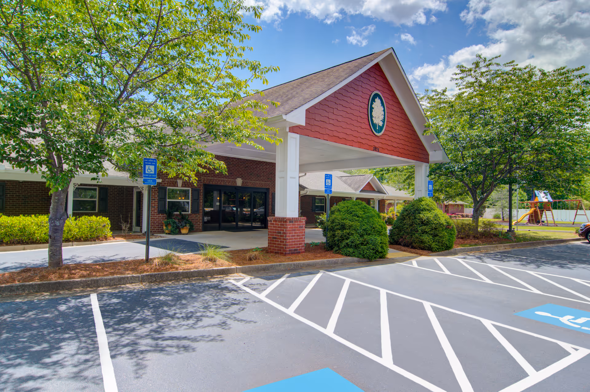 Entrance of a senior living facility with a covered drop-off area supported by white columns and a red gable featuring a tree emblem. There are several handicap parking spaces in front, surrounded by green trees and bushes under a partly cloudy sky.