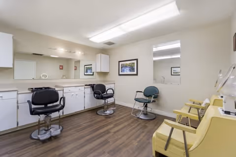 Interior view of a salon area in a senior living facility with three salon chairs, a large mirror on the wall, white cabinets, framed pictures, and yellow waiting chairs along one wall. The room has wood flooring and bright overhead lighting.