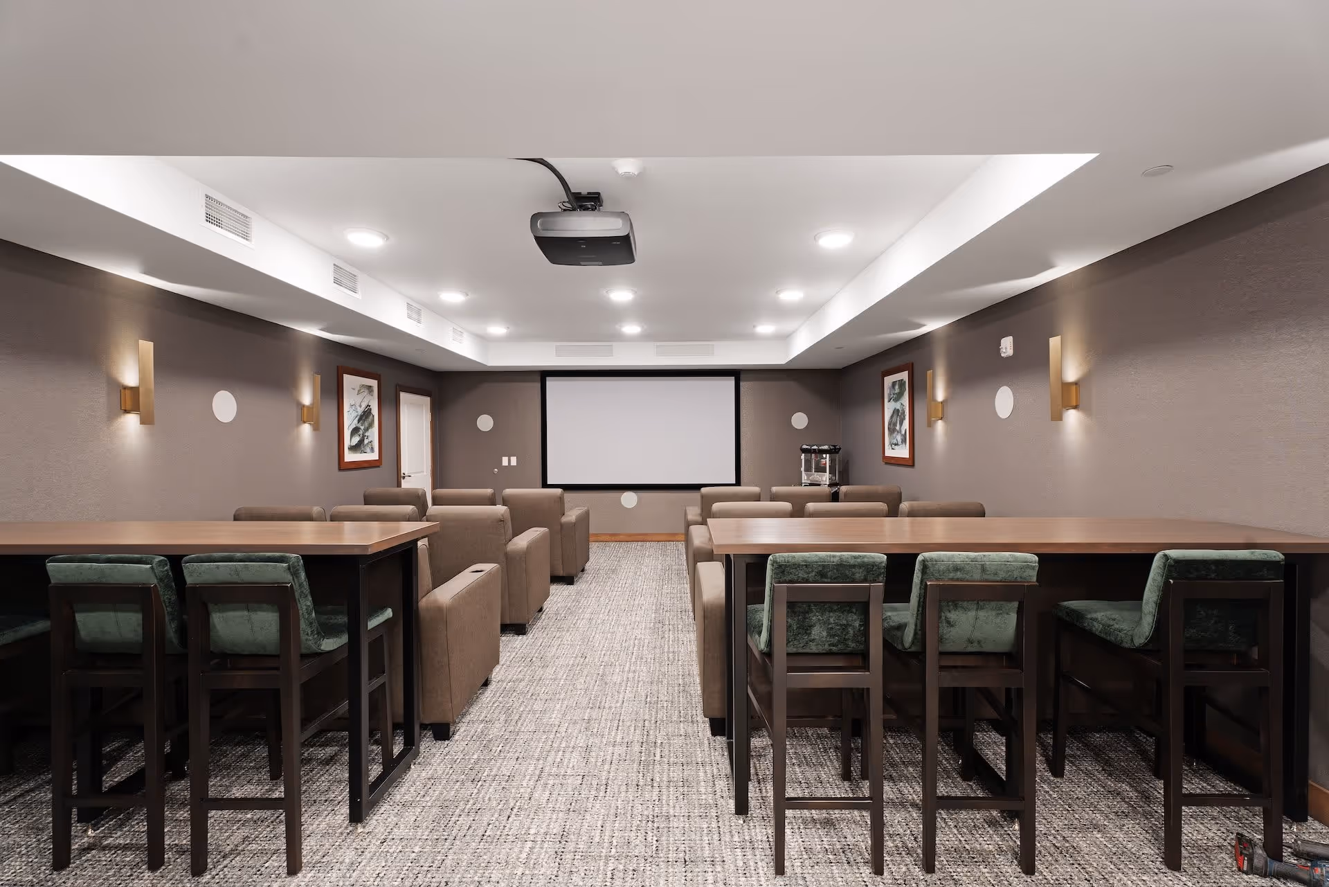 A modern media room with a large projector screen at the front, surrounded by brown armchairs arranged in rows. There are two high tables with green cushioned chairs on either side of the room. The walls are decorated with framed artwork and wall-mounted lights, and the ceiling has recessed lighting and a ceiling-mounted projector.