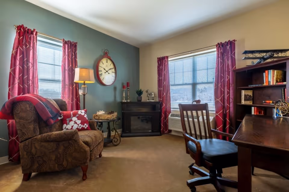 A cozy living room with a patterned armchair draped with a red and blue blanket and a floral pillow, a floor lamp, a round side table, a wall clock, a small black electric fireplace, two windows with red curtains, a wooden desk with a rolling chair, and a bookshelf with books and decorative items.