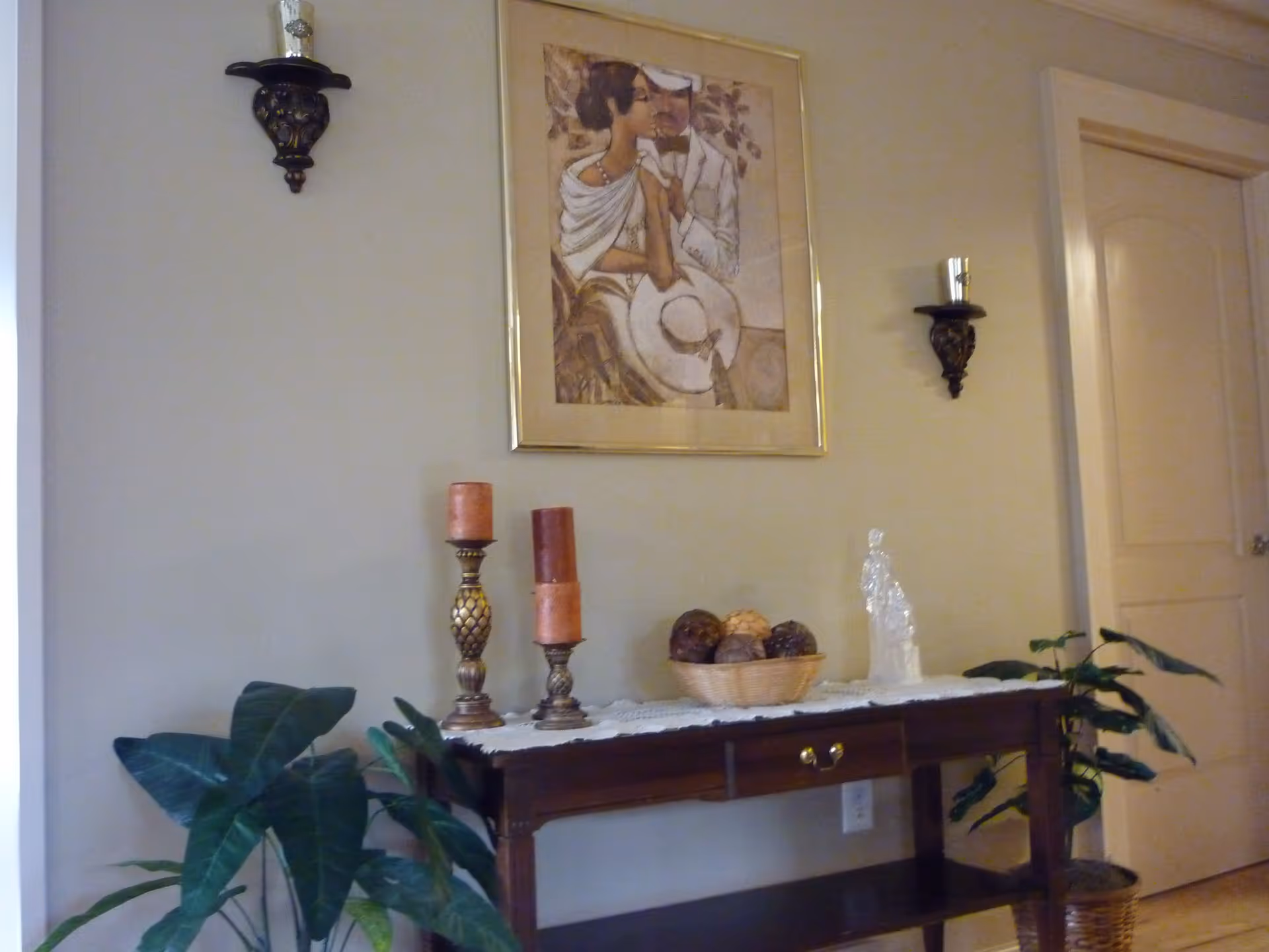 Interior hallway with a wooden console table holding candles, a decorative bowl and a glass figurine, flanked by potted plants and a framed print with wall sconces.