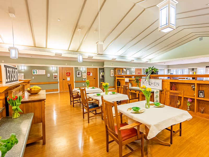 A bright and spacious dining area with wooden floors and tables covered with white tablecloths. Each table has green bowls, plates, and vases with yellow tulips. Wooden chairs surround the tables, and there are decorative elements like framed pictures and plants. The ceiling has hanging lantern-style lights, and a sign labeled 'KITCHEN' is visible on a wooden partition.