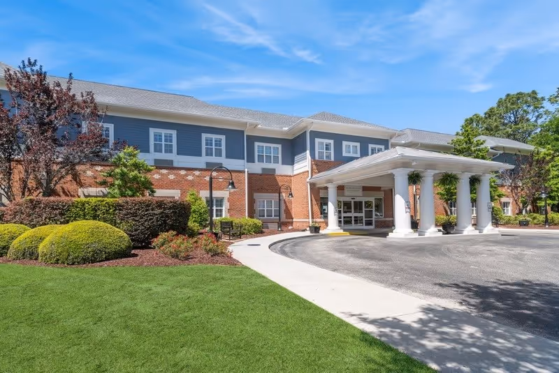 Exterior view of a two-story senior living facility building with a covered entrance supported by white columns. The building features a combination of red brick and blue siding with multiple windows. There is a well-maintained lawn, landscaped bushes, and trees surrounding the entrance area under a clear blue sky.