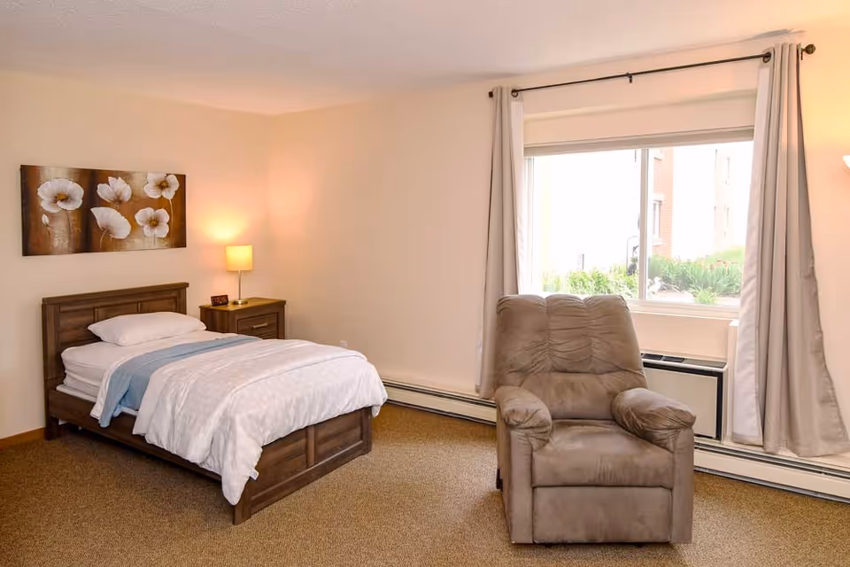A cozy bedroom with a single wooden bed made with white bedding and a light blue throw. Next to the bed is a wooden nightstand with a lamp and a small clock. A large window with beige curtains lets in natural light, and a comfortable gray armchair is positioned near the window. The walls are painted light beige, and there is a floral painting hanging above the bed.