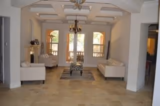 A spacious living room with beige tiled flooring, two white sofas facing each other, a patterned armchair in the center, a chandelier hanging from the ceiling, and large windows with arched frames letting in natural light.