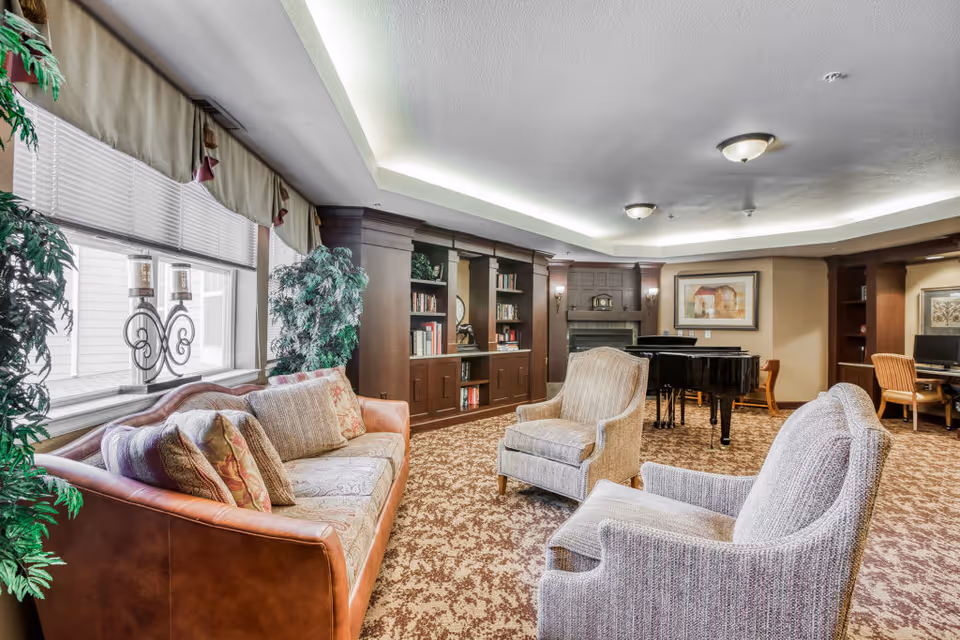 A cozy living room area in an assisted living facility featuring a brown leather sofa with patterned cushions, two upholstered armchairs, a grand piano, built-in dark wood bookshelves, a fireplace, and a desk with a computer in the background. The room has a patterned carpet, large windows with blinds and valances, and soft ceiling lighting.