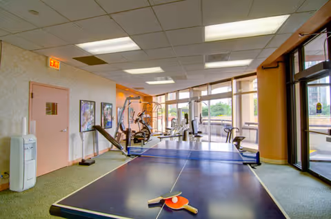 Indoor recreational room with a ping pong table in the foreground, exercise equipment including a stationary bike and weight machine in the background, large windows letting in natural light, and a water cooler near a closed door with an exit sign above it.