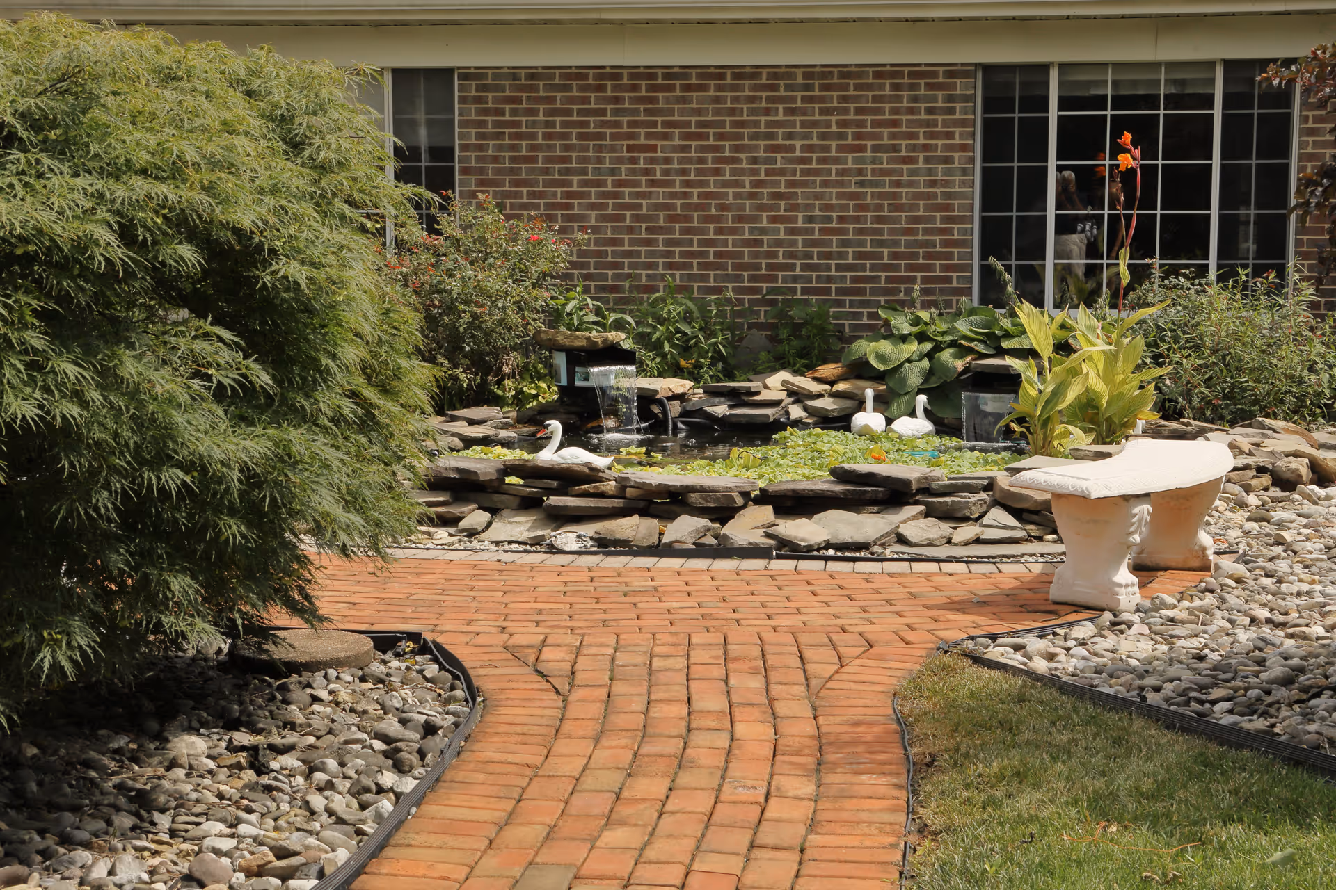 Outdoor garden area with a brick pathway leading to a small pond surrounded by rocks and plants. There is a white stone bench on the right side and a brick building with windows in the background.