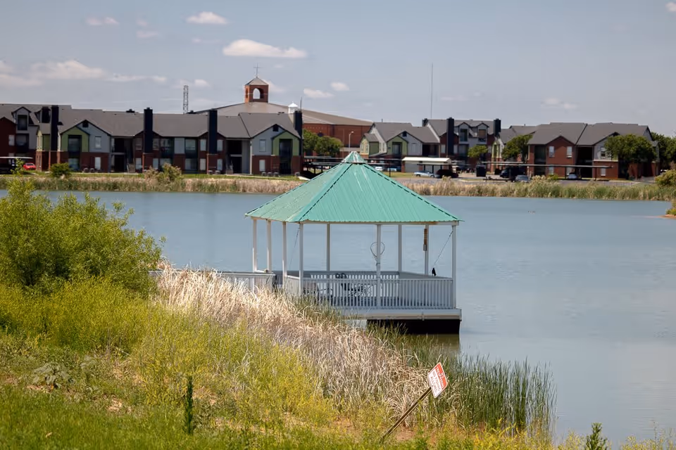 A gazebo with a green roof situated on the edge of a lake with tall grass and shrubs in the foreground. In the background, there are multiple two-story residential buildings and a church with a cross on top under a partly cloudy sky.