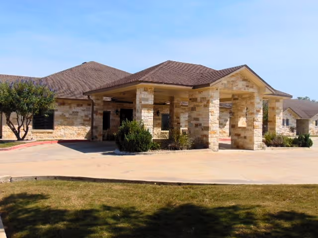 Exterior view of a single-story nursing and rehab center building with stone walls and a brown shingled roof. The entrance features a covered driveway supported by stone pillars, with some bushes and trees around the building under a clear blue sky.