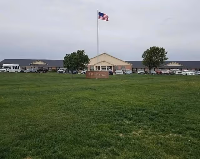 Large grassy lawn leading to the front of the Prairie House senior living building with an American flag on a tall flagpole and a sign.