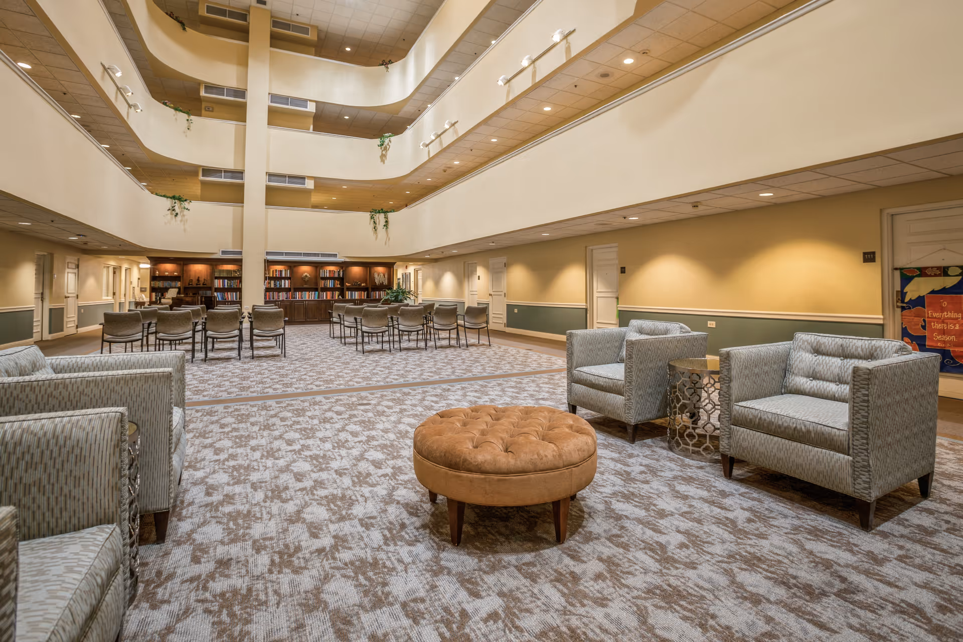 Spacious and well-lit common area in a senior living facility with multiple seating arrangements including armchairs and a round ottoman. The area features a high ceiling with multiple floors visible, a carpeted floor, and a bookshelf filled with books at the far end. The walls are painted in warm tones with some plants hanging from the upper floors.