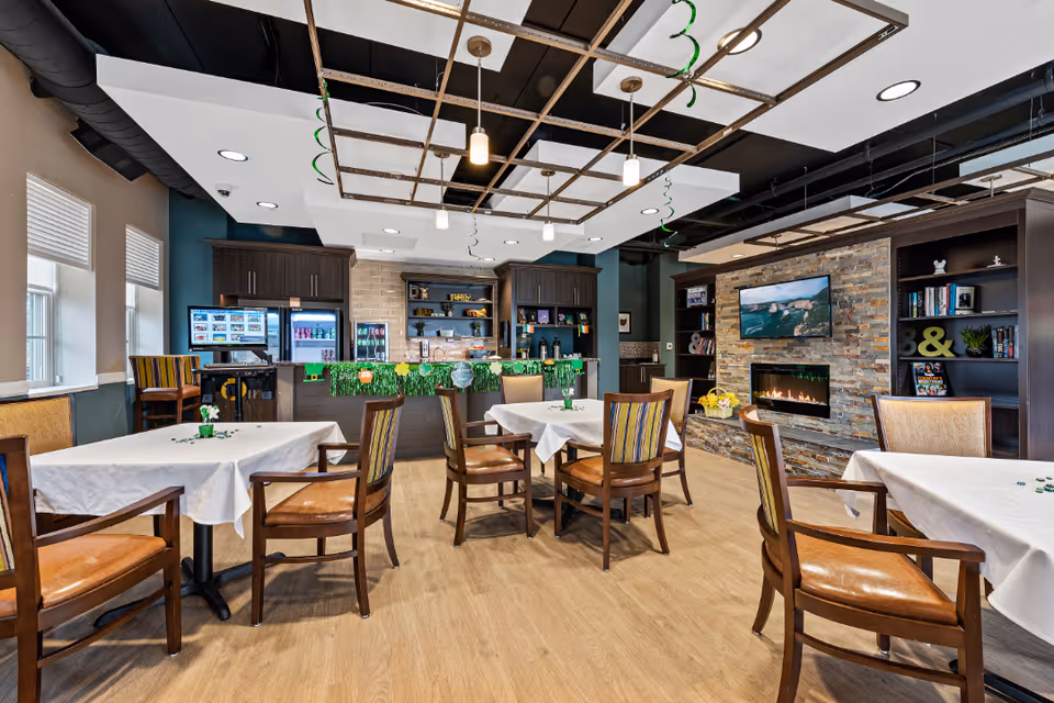 Bright communal dining area with tables and chairs, a service counter/kitchenette, and a fireplace with a wall-mounted TV.