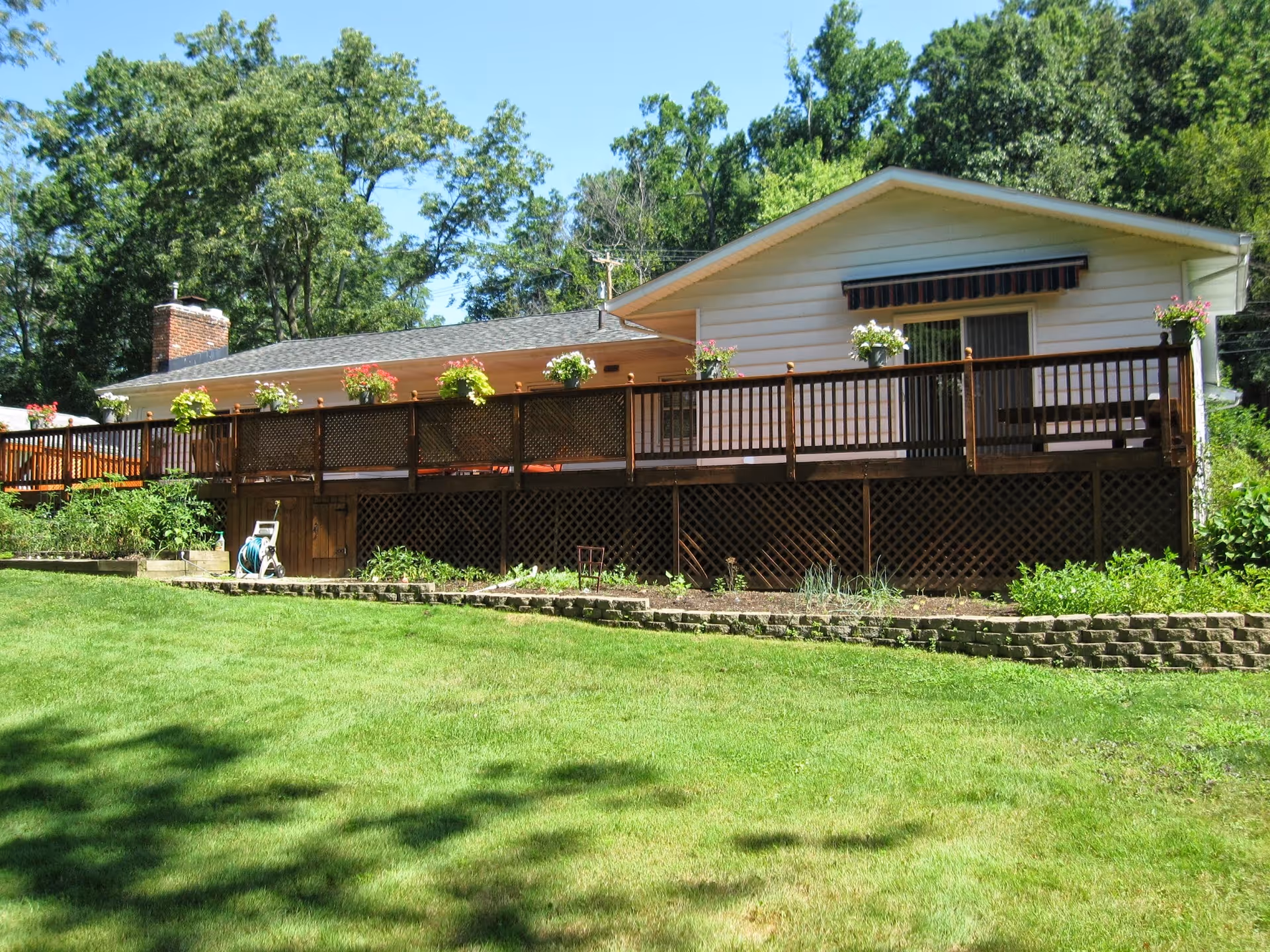 A single-story house with a large wooden deck adorned with flower pots. The house is surrounded by green grass and trees under a clear blue sky.
