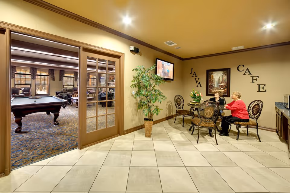 Small indoor café seating area with two women talking at a table near a potted plant and a wall-mounted TV, with a view into a game room containing a pool table.