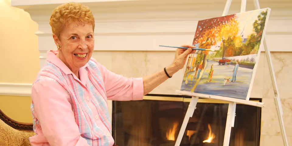 An elderly woman with short curly hair wearing a pink shirt and a pastel plaid vest is sitting in a chair and painting on a canvas set on an easel. The painting depicts a colorful outdoor scene with people and trees. Behind her is a lit fireplace with a beige marble surround.