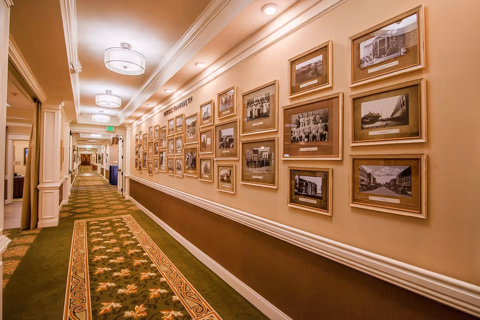 A long, well-lit hallway with a green and beige patterned carpet. The right wall is decorated with numerous framed black and white photographs arranged in a gallery style. The ceiling has modern light fixtures, and the left side of the hallway has doorways leading to other rooms.