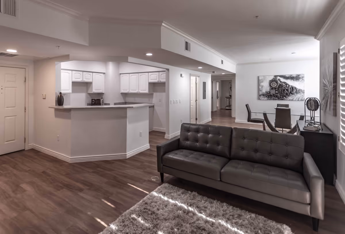 A modern living area with a gray leather sofa on a shaggy rug, a glass dining table with chairs, and a kitchen with white cabinets and a breakfast bar. The room has wood flooring and white walls, with a black and white train painting on the wall near the dining area.