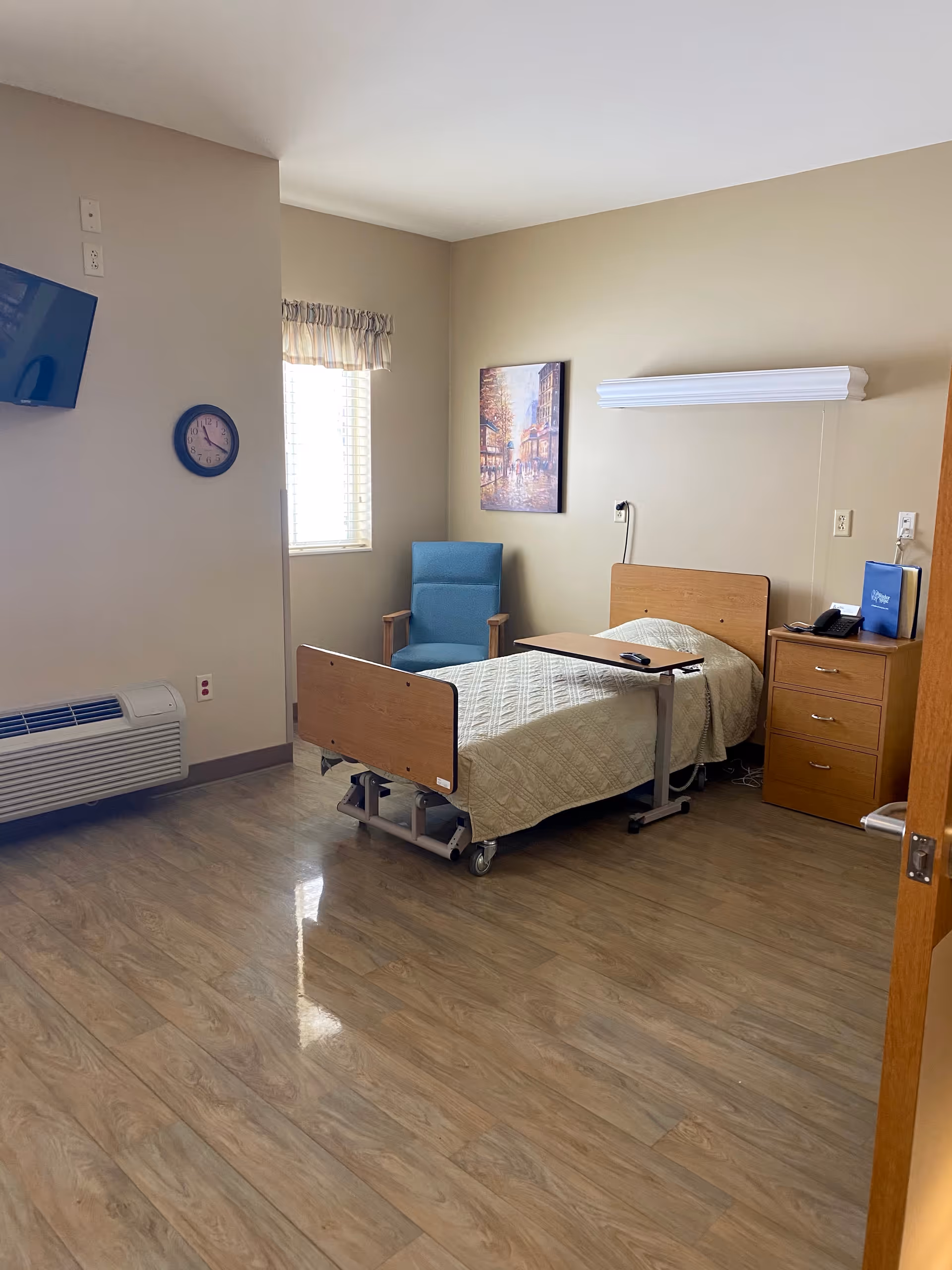 A simple, clean bedroom in a senior living facility with a single hospital-style bed covered with a beige quilt, a blue upholstered chair, a wooden nightstand with drawers, a wall-mounted TV, a clock, and a window with blinds and a valance. The floor is wood-patterned, and there is a piece of artwork on the wall above the chair.