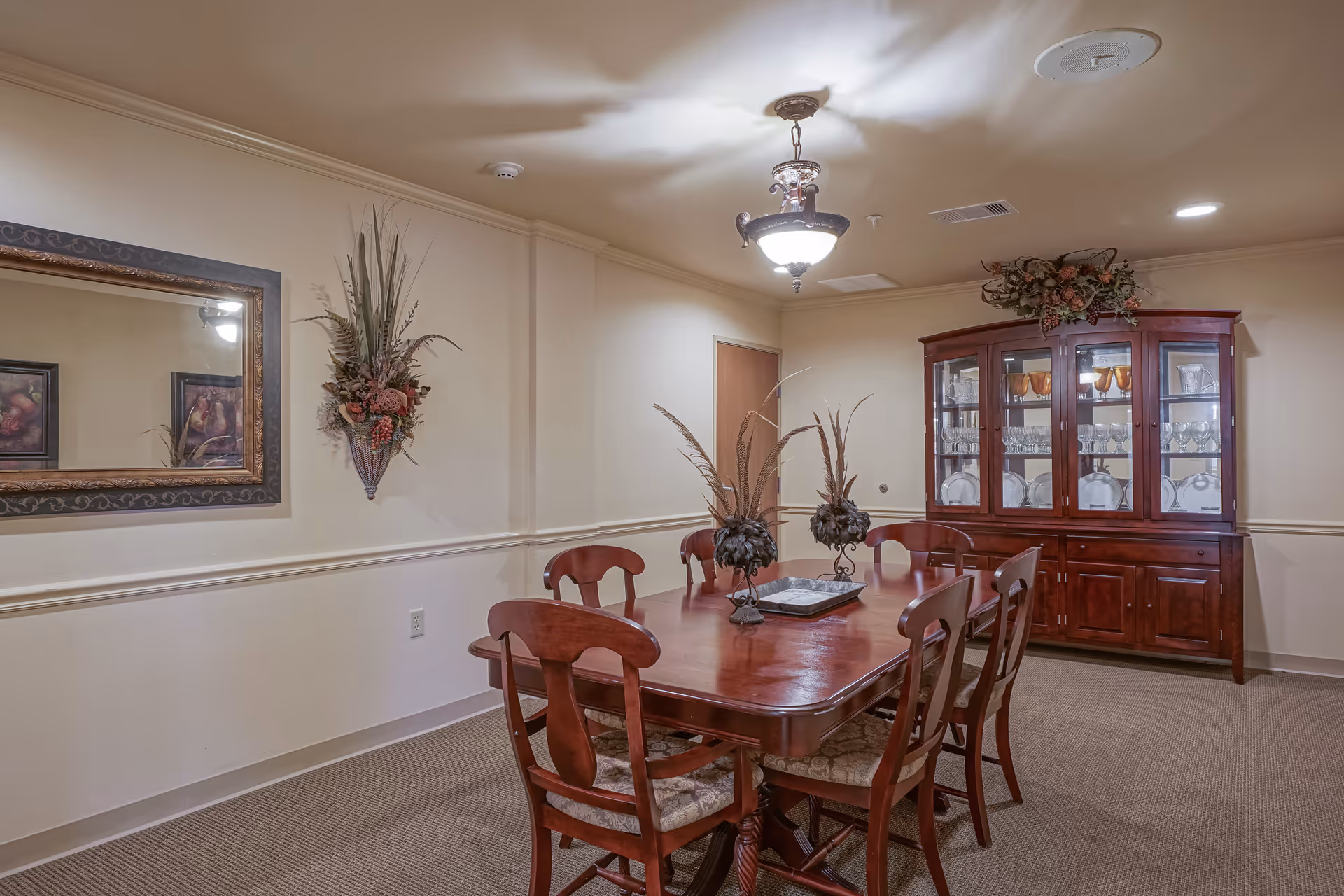 A dining room with a wooden dining table and six matching chairs. The table has decorative centerpieces with tall feathers. On the right side, there is a wooden china cabinet displaying glassware and plates. The walls are beige with a framed mirror and a floral wall decoration. The room is lit by a ceiling light fixture.