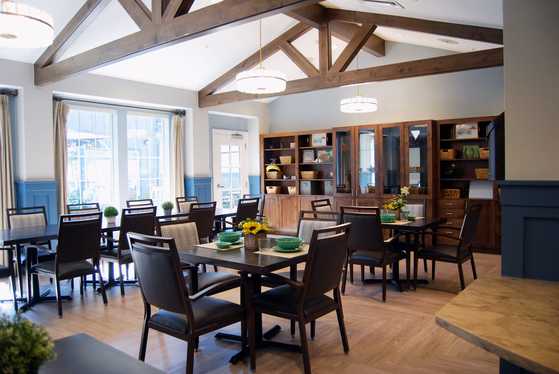 A bright and spacious dining room with wooden tables and chairs, decorated with small plants and flowers. The room features large windows with curtains, wooden ceiling beams, and built-in wooden cabinets along the back wall.