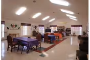 A spacious, well-lit common area in a senior living facility with several tables covered with purple and orange tablecloths, surrounded by chairs. The room has high ceilings with skylights, windows with curtains, and various decorations on the walls. There are bookshelves and seating areas along the walls.