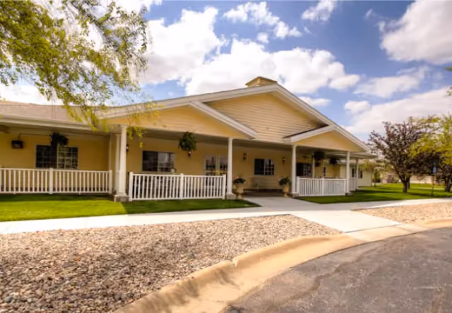 Single-story yellow senior living building with a covered white-railed porch, hanging plants, and a front walkway under a partly cloudy sky.