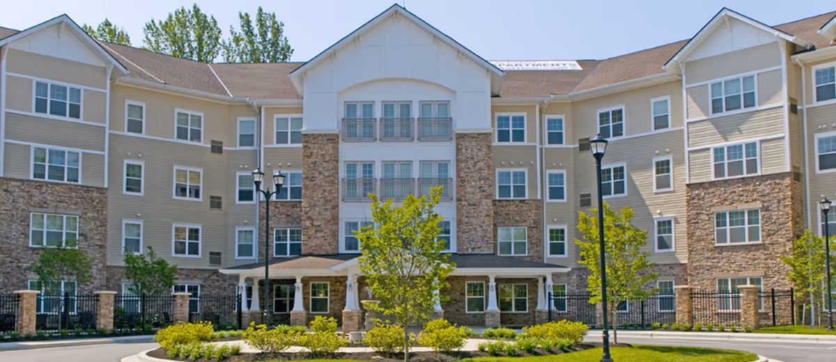 Front exterior view of a multi-story senior living facility with beige siding and stone accents, surrounded by landscaped greenery and trees under a clear sky.
