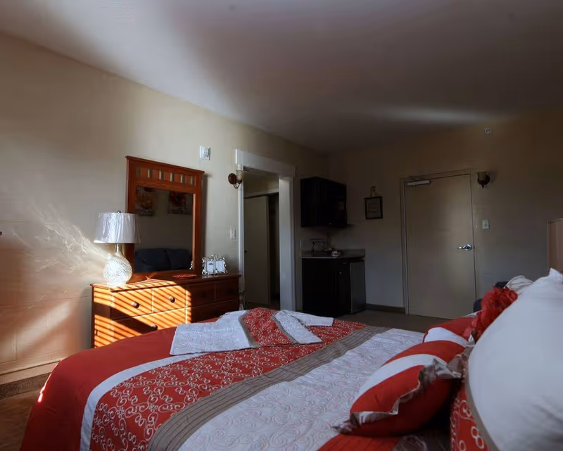 A bedroom in a senior living facility with a bed covered in red and white patterned bedding and pillows. A wooden dresser with a mirror and a lamp is against the wall, with sunlight casting shadows through blinds. There is a small kitchenette area with a mini fridge and cabinets, and a closed door in the background.