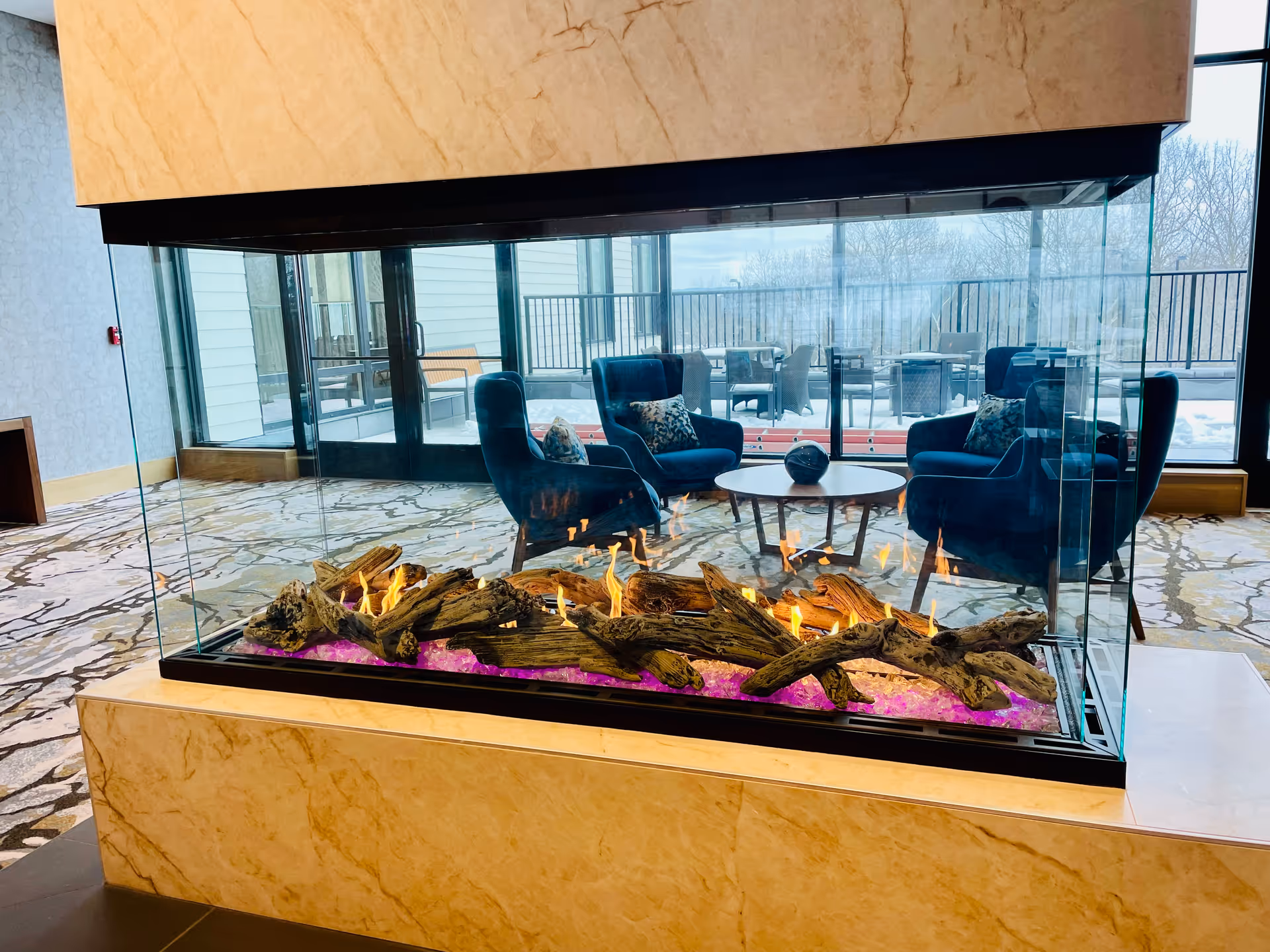 A modern indoor fireplace with glass panels on all sides, containing burning logs and purple decorative stones. Behind the fireplace, there are blue upholstered armchairs with patterned pillows arranged around a round wooden coffee table. Large windows and glass doors reveal an outdoor patio area with tables and chairs, and some snow is visible outside.