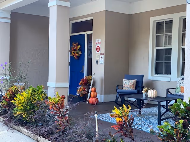 Covered porch area with a blue door decorated with a fall-themed wreath. There are stacked pumpkins and a small table with two chairs, each with decorative pillows. The porch is surrounded by colorful plants and shrubs.