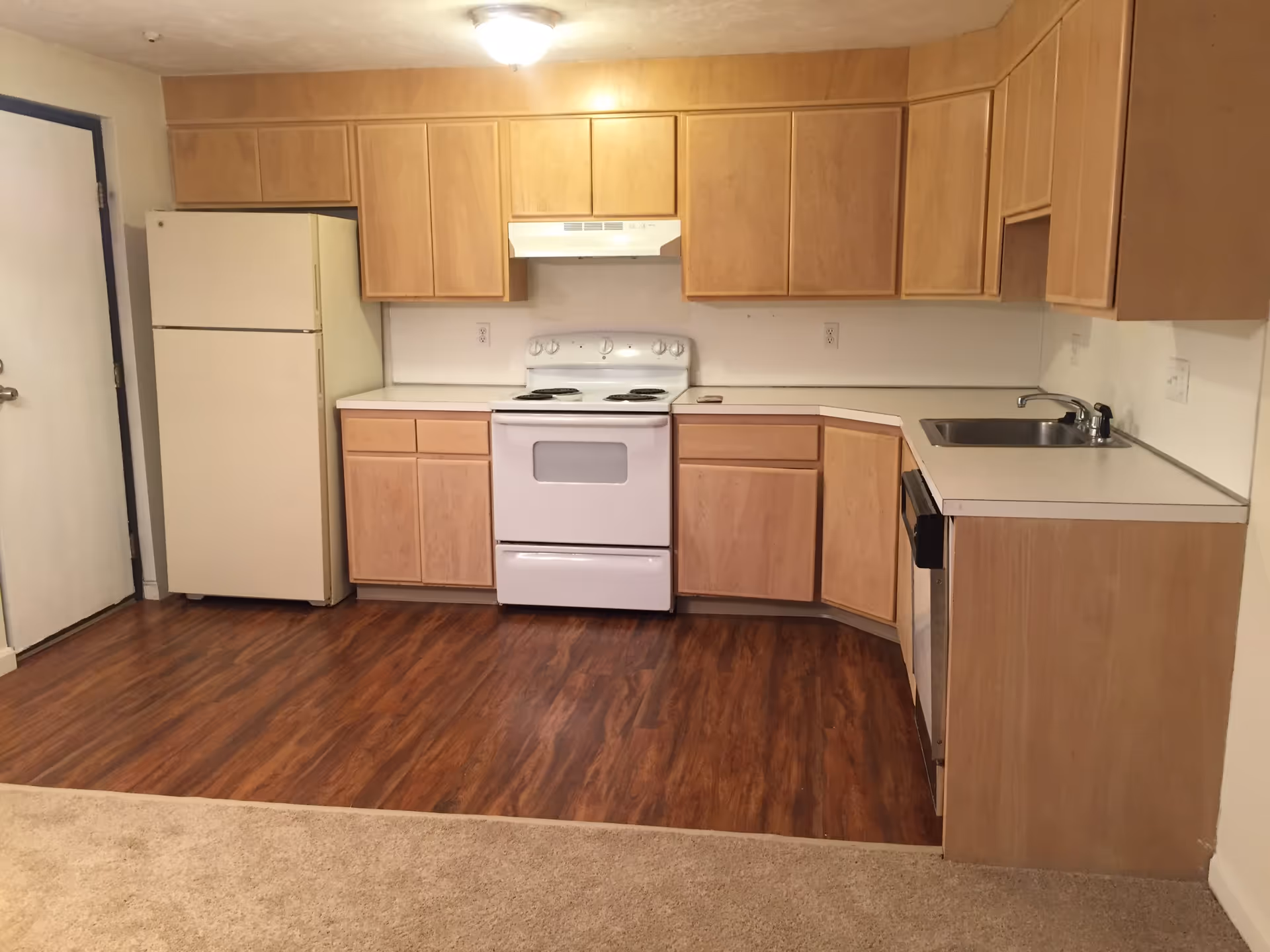 A kitchen area with light wood cabinets, a white refrigerator, a white electric stove with an overhead vent, a stainless steel sink, and a dishwasher. The floor is a combination of wood and carpet, and there is a closed door to the left.