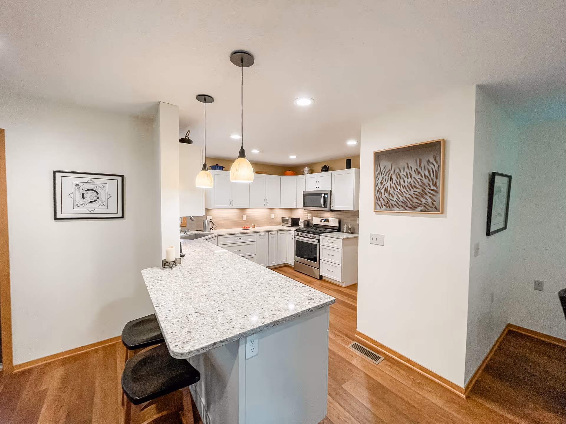 Bright and modern kitchen with white cabinets, granite countertops, and wooden flooring. Two pendant lights hang over a breakfast bar with two black stools. Stainless steel appliances including a stove and microwave are visible. Artwork is hung on the walls.