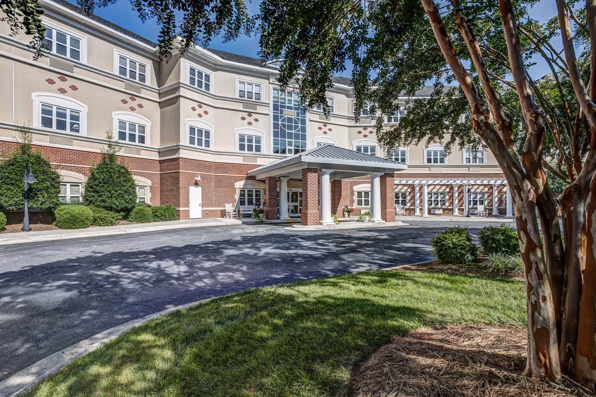 Front exterior of a multi-story senior living facility with a covered drop-off entrance, curved driveway, and landscaped trees.