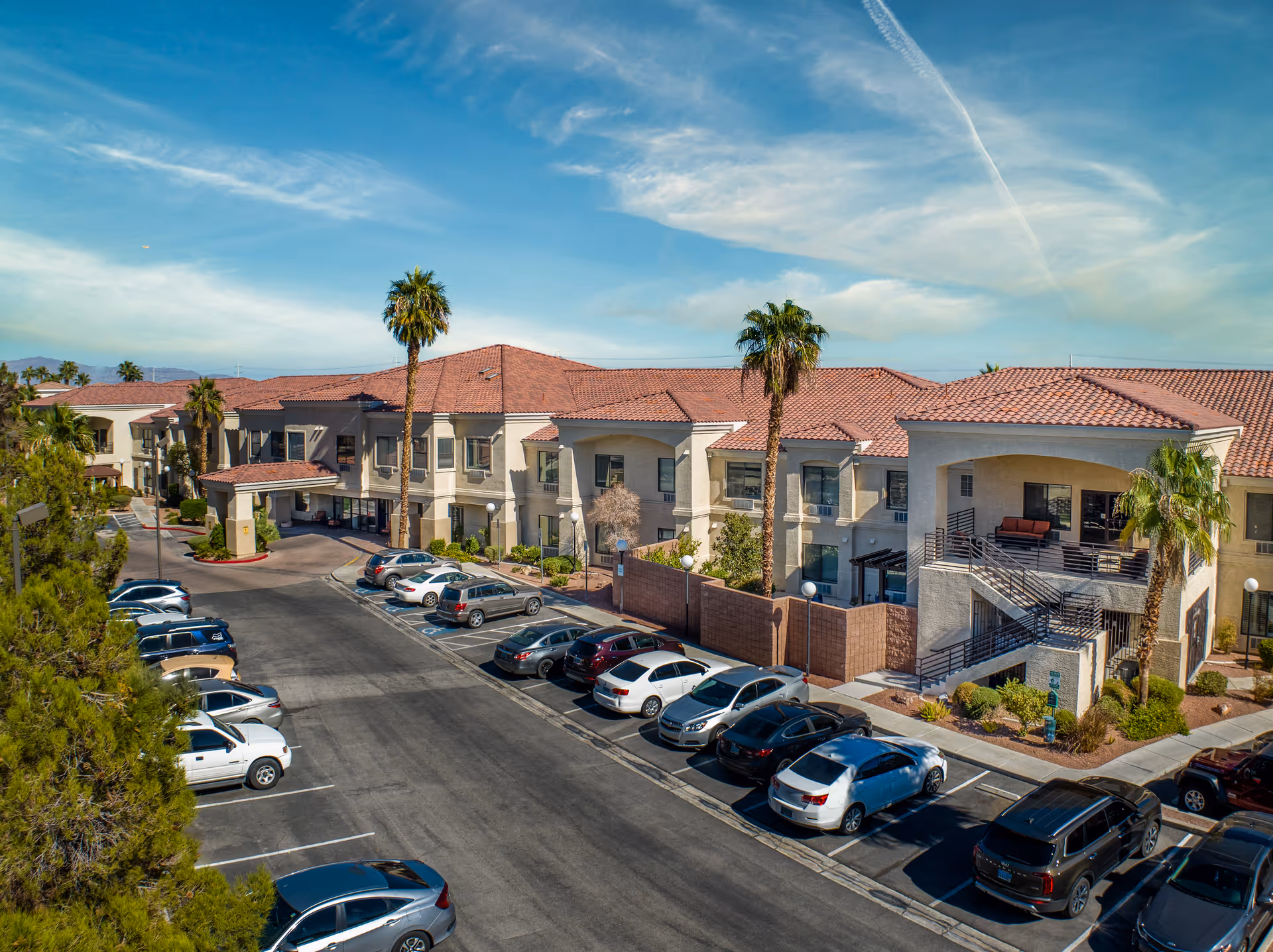 Front exterior of a two-story senior living building with red tile roofs, palm trees, balconies, and a parking lot.